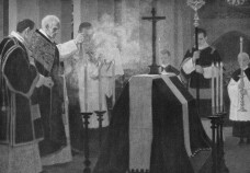 Totenmesse: Der Priester steht vor dem Sarg, der mit einem schwarzen Tuch verhüllt ist und auf dem ein Kreuz aufgestellt ist; der Priester inzensiert mit Weihrauch den Sarg; Ministranten stehen ebenfalls dabei
