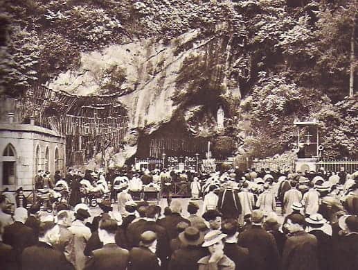 Pilger vor der Grotte in Lourdes: man sieht die vielen Krücken der Geheilten an der Wand des Berges, die Muttergottes-Statue sowie den heiligen Ort in der Grotte, durch die Absperrung geschützt; vor dieser befinden sich die Pilger und Kranken, die um Heilung bitten