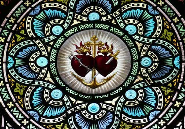 Ein Glasfenster in Montserrat: in der Mitte des ornamentalen Fensters ist ein goldener Anker mit einem Kreuz zu sehen sowie zwei Herzen, eines mit Dornen umrankt, das andere mit einem Schwert durchbohrt, die Herzen Jesu und Mariens