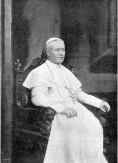 Pius X. in weißer Papstkleidung sitzt auf dem päpstlichen Stuhl, die Arme auf den Stuhllehnen, im Gesicht ist ein leichtes Lächeln zu sehen